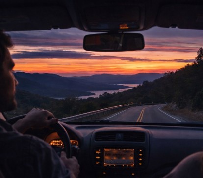 View from the front passenger seat of a car driving down a winding road at sunset. The dashboard and steering wheel are visible, and the sky outside glows with vivid orange, pink, and purple hues over distant hills and water.